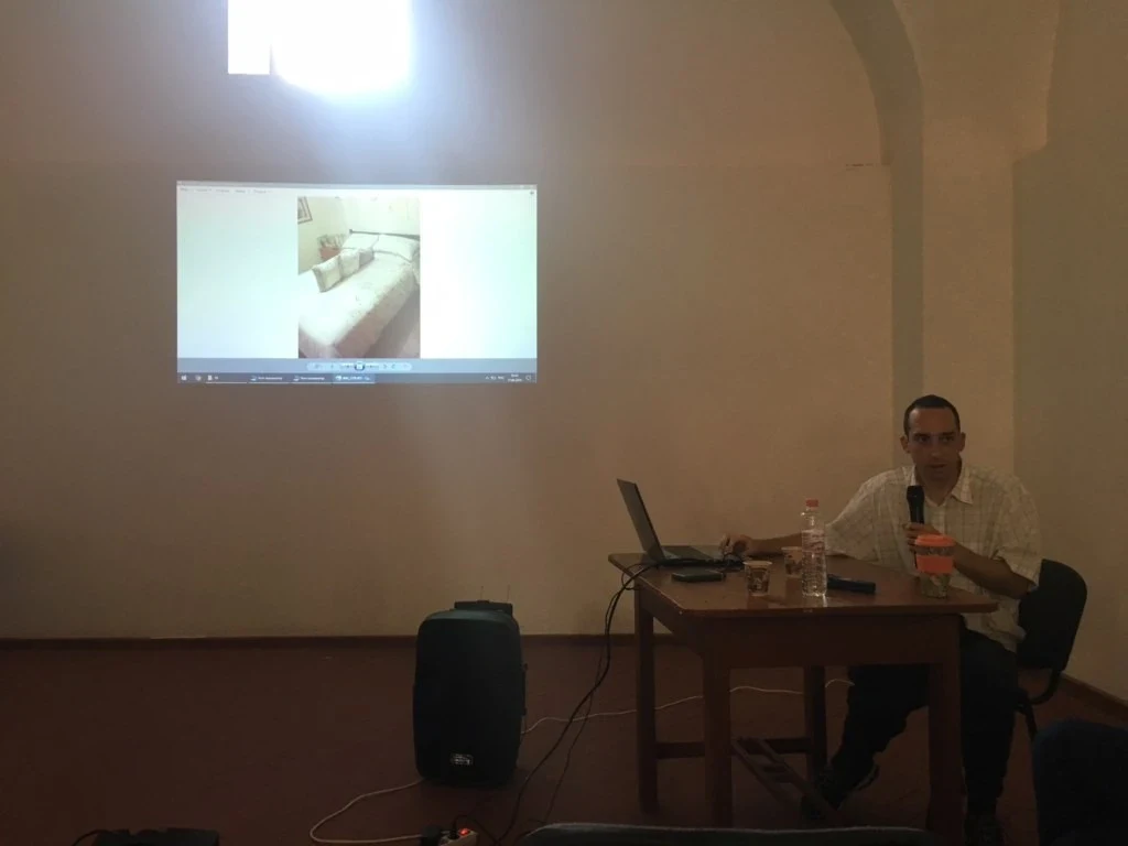 A speaker seated at a table beneath two projected images.