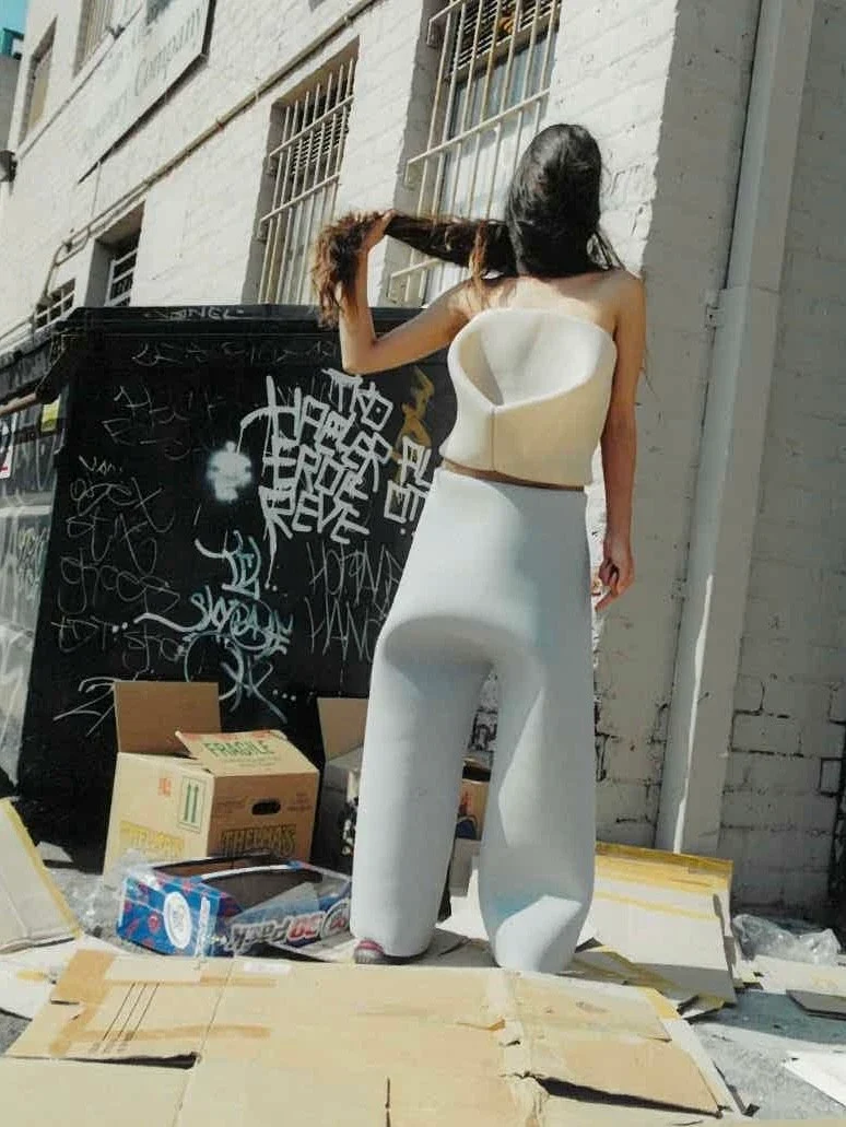 A person standing in a studio wearing a white cropped top and pale trousers.