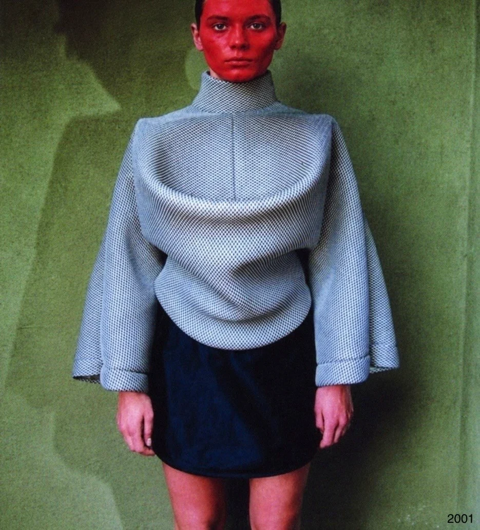 A person in a gray sweater with a large draped front and a red face covering.